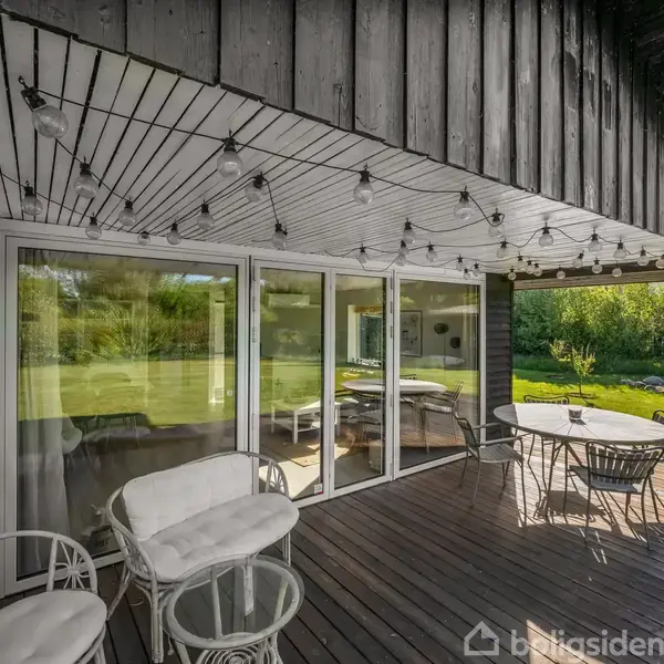 Terrasse med hvide stole og bord, omgivet af glasvægge. Lyskæder hænger fra loftet. Grønne buske og græsplæne i baggrunden.