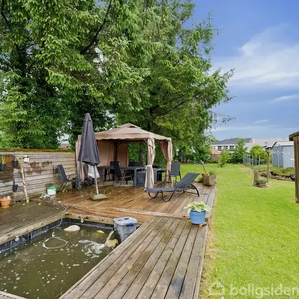 Terrasse med trædæk omkranser en lille dam, der indeholder liljer. En pavillon med møbler står nær, omgivet af græsplæne og træer i en have.