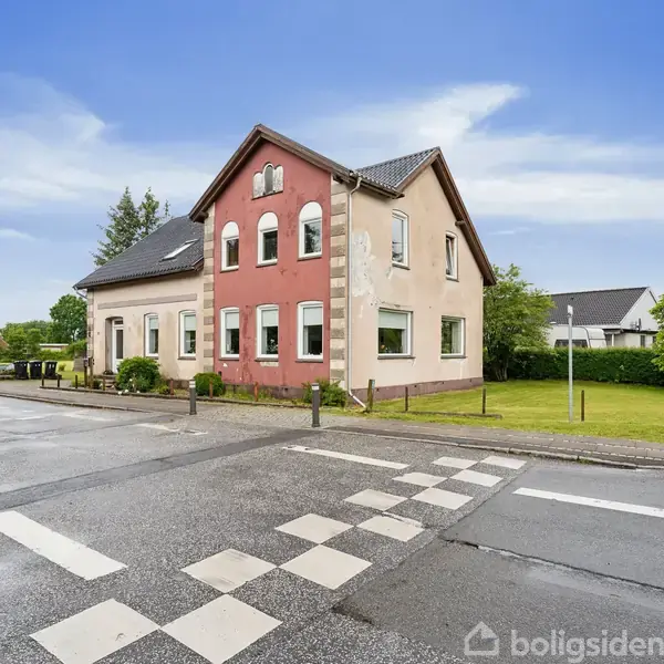Et to-etagers hus står ved en vejkryds. Det har en rød facade i midten og en beige facade i siderne. Det grønne område omgiver huset.
