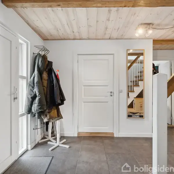 Jakker hænger på en frakkestativ ved siden af en hvid dør, i en indgang med lyse trægulve, en trappe og en spejl langs væggen.