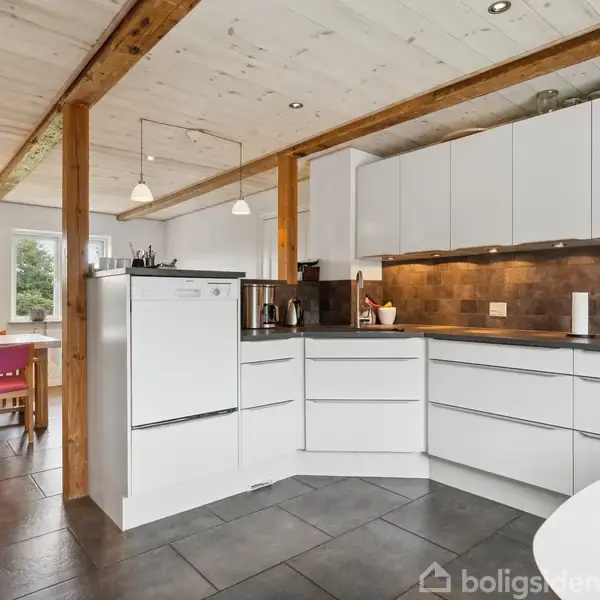 Hvidt køkken med moderne skabe og skuffer, kaffemaskine og stearinlys på bordpladen. Træbjælker i loftet. Til venstre ses spiseplads med lyse træmøbler og lyserøde hynder.