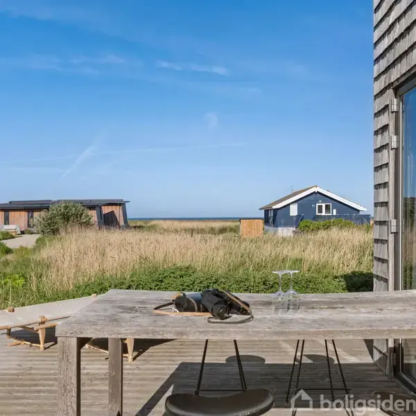 Træbord med stole står på en terrasse foran et kystnært sommerhus. Baggrunden viser græsarealer, to andre små huse og havet i horisonten under en klar blå himmel.