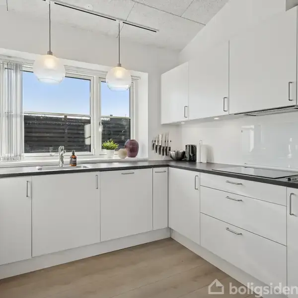 Et moderne køkken med hvide skabe og skuffer. To hængende lamper lyser rummet op. Vindue med udsigt og sort bordplade med indbygget kogeplade og knivholder ved siden.