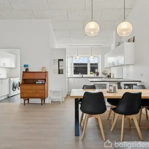Spisebord med sorte stole står i et lyst køkken-alrum. Bordet er dækket med en kande. Vægskabe og apparater fylder køkkenet. Døren til venstre fører til et vaskerum.