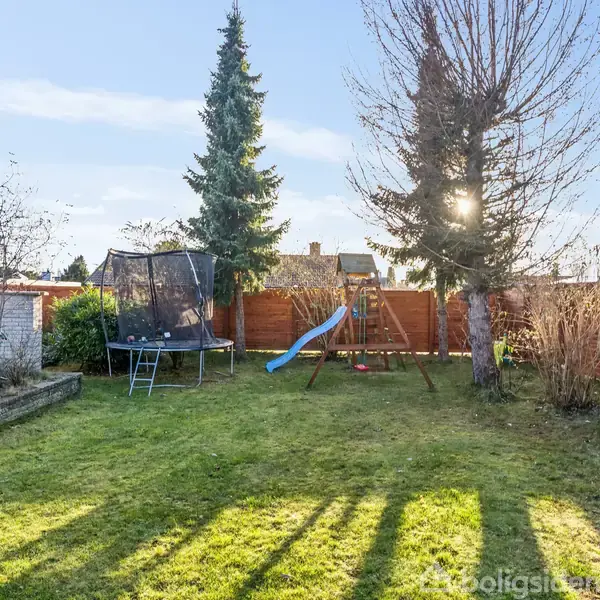 En have med en trampolin og en blå rutsjebane på en legestativ, omgivet af grønne plæner og store træer mod en klar, solrig himmel.