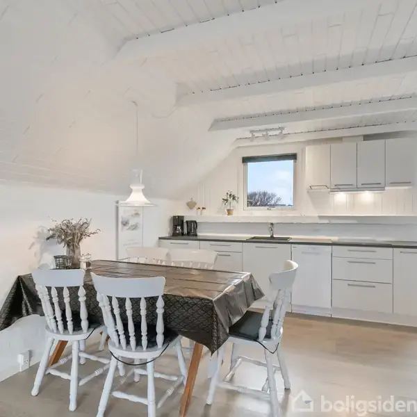Et spisebord med fire hvide stole står i et moderne køkken med hvide skabe. Bordet har en sort dug, og en lampe hænger over det. En stor dør åbner ud mod haven.