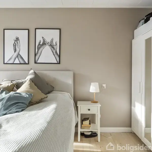 En seng står midt i et lyst soveværelse med puder og sengetæppe, flankeret af natbord med lampe. Væggen har to indrammede kunstbilleder. En hvid garderobe er til højre.