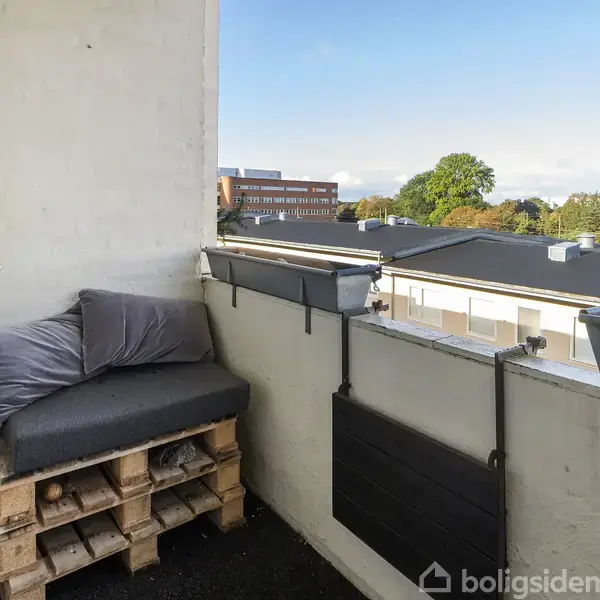 En lille balkon med en sofa lavet af paller og grå puder, står ud mod en udsigt over hustage og træer i fjernsynet. Blomsterkasser hænger på rækværket.