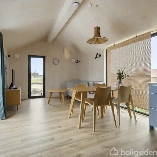 Et moderne, lyst rum med spisebord og stole. Et tv står ved siden af en stor glasdør, der åbner til en have. Trægulv og et blåt køkken ses til højre.