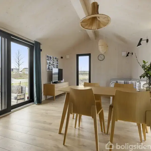 Spisebord med fire stole står i en lys stue med trægulv. Store vinduer til venstre giver udsigt til en udendørs terrasse. En vase med blomster pryder bordet.