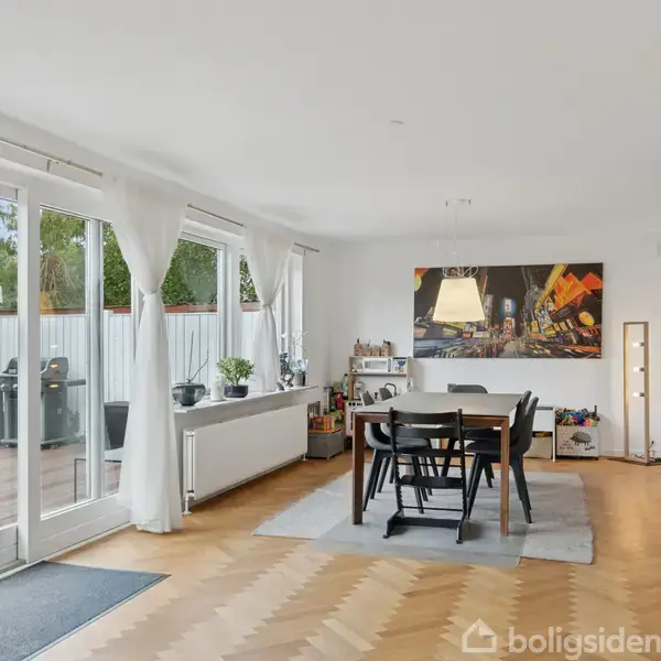 Spisebord med seks stole står i et lyst rum med trægulv og vægkunst. Store vinduer åbner til en terrasse med udendørs siddepladser, omgivet af planter.