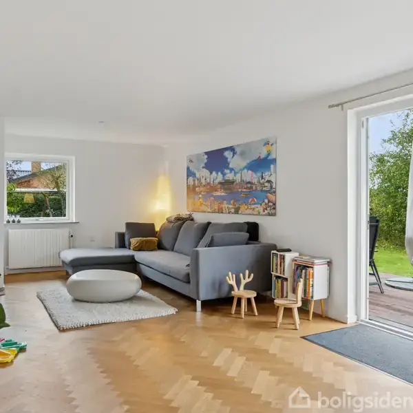 Stue med grå sofa og sofabord står ved siden af en stor glasdør, der åbner ud til en terrasse med grøn udsigt. Et maleri hænger over sofaen, omgivet af legetøj.