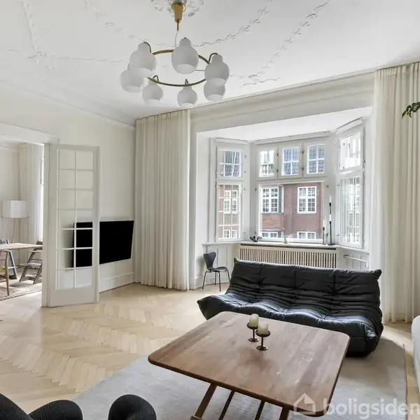 En stue med en sort sofa vender mod et træbord, indrettet med lyse hvide vægge og en stor vindueskarnap. Til venstre er en åben skydedør til en spisestue.