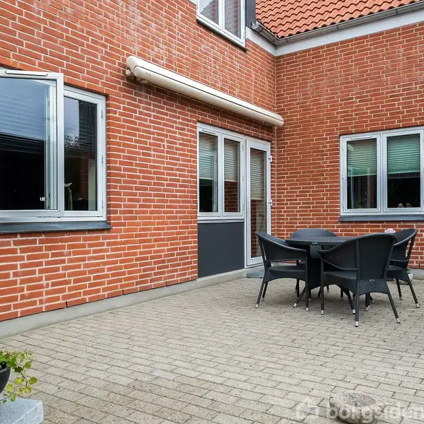 Rundt havebord med fire stole står på en flisebelagt terrasse. Omgivet af en rød murstensbygning med vinduer og glasdør, der fører ind i huset.