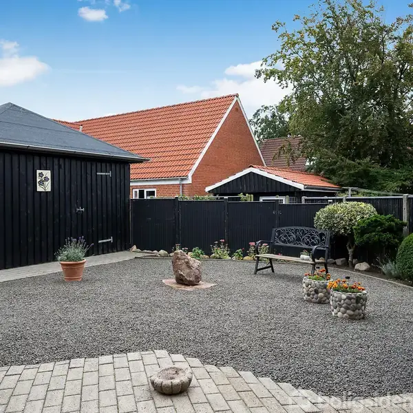 En grusgårdsplads med en sortmalet skur og bænk omgivet af buske og potteplanter. I baggrunden ses rødt tag og et hegn, der skærmer området.