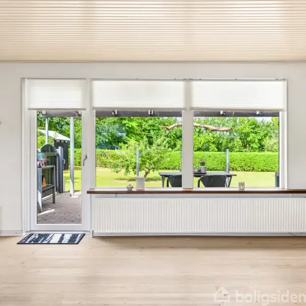 En stol står ved siden af en åben glasdør, der fører ud til en terrasse og grøn have. Inde i rummet ses et langt radiatorpanel under vinduerne.