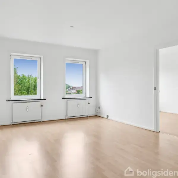 Tomt værelse med trægulv og to vinduer, der lukker naturligt lys ind. En hvid dør fører ind til et tilstødende rum. Væggene er hvide. Udsigt til grønt udenfor.