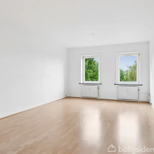 Et lyst og tomt værelse med trægulv, to store vinduer hvorigennem grønne træer er synlige. Rummet har hvide vægge og lediger plads.