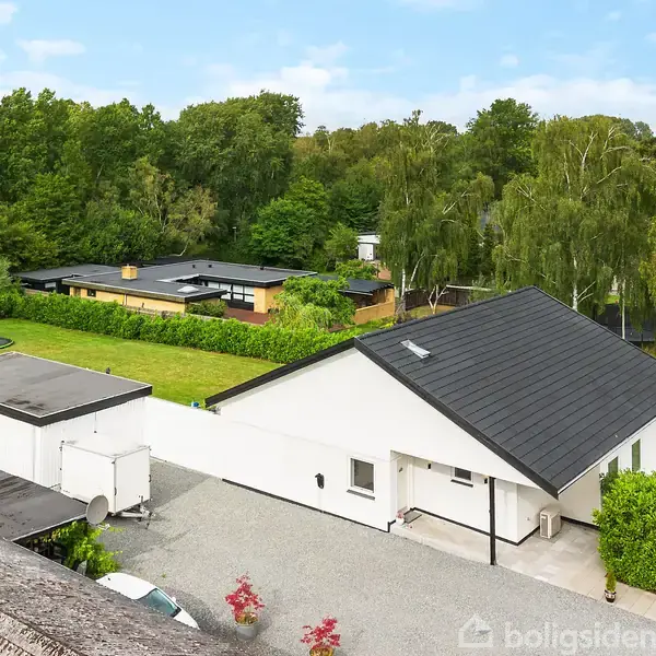 En hvid enetages villa med sort tag står på en grusbelagt indkørsel, omgivet af grønne buske og træer i en skovklædt baggrund. Legeredskaber ses i en tilstødende have.