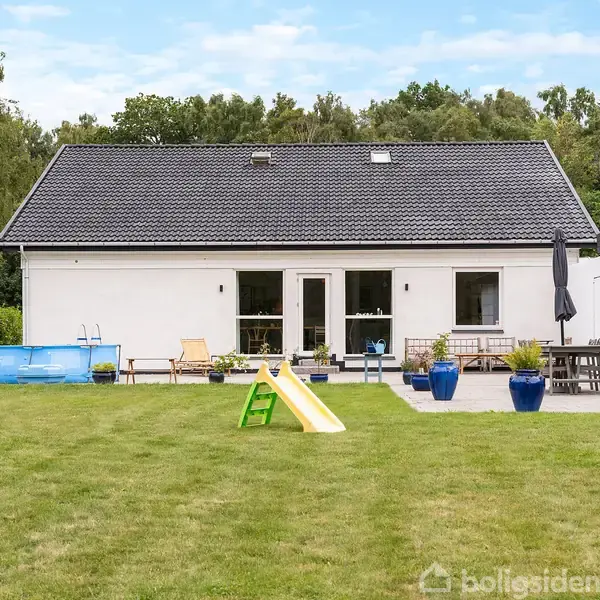 Et hvidt hus med sort tag står i en have. En gul-rutschebane og et lille blåt badebassin er på græsplænen. Terrassen har møbler og blå krukker.