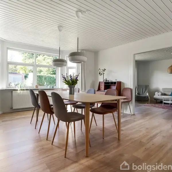 En spisebord med seks stole står i et lyst rum med parketgulv. Store vinduer giver udsigt til en frodig have. Tilstødende stue ses med sofa og dekorationer.