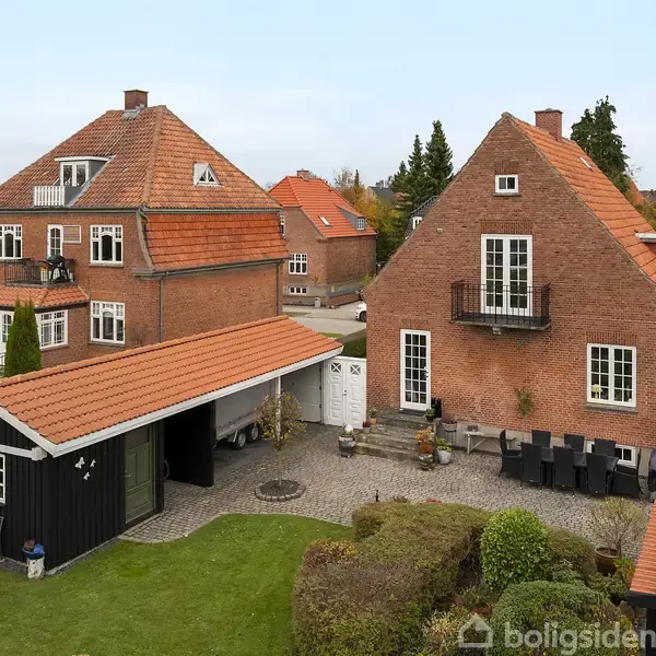 Rødt murstenshus med sort carport i forhaven, omgivet af en grøn have og andre lignende huse med røde tage. Der ses også en terrasse med havemøbler.