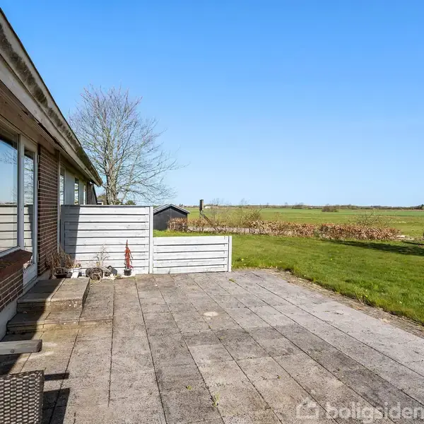 En stor terrasse med fliser set fra et hus med store vinduer. Terrassen er omgivet af en græsplæne og åbent landskab under en klar blå himmel.
