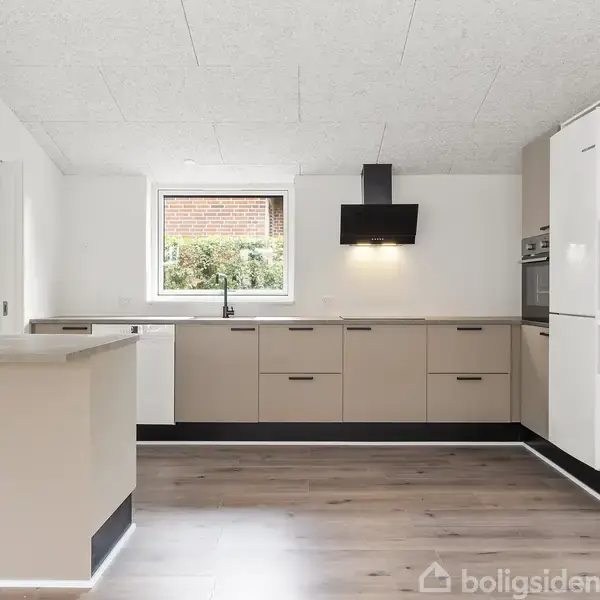 En moderne køkkenindretning med beige skabe og indbygget ovn står i midten, omkranset af lyse vægge og trægulv. En emhætte hænger over komfuret, og et vindue giver naturligt lys.