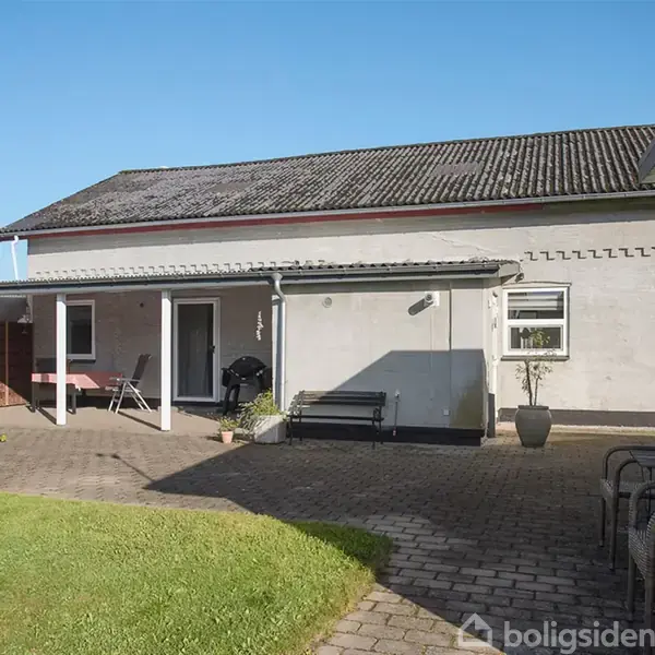 Et hus med en flisebelagt terrasse, hvor grill og havemøbler står. Grønne omgivelser inkluderer græsplæne og buske, og en skyggefuld veranda tilbyder siddepladser.