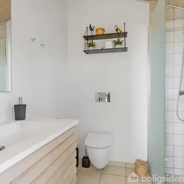 Håndvask står i et moderne badeværelse med hvidt interiør. Ved siden af er der et toilet, hylder med planter og en bruser bag glasvægge. Rummet har lyse fliser og træloft.