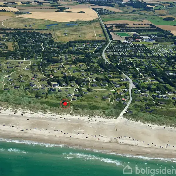 Kyststrækning med huse fordelt i grønne områder tæt ved stranden. Biler og mennesker ses langs vandkanten. Omkringliggende landskab består af marker og skove.