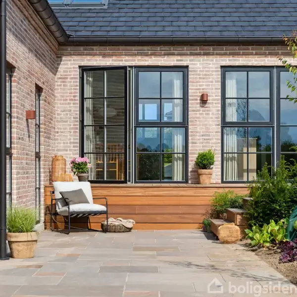 En moderne terrasse har en komfortabel stol med puder placeret ved siden af blomsterkrukker. Store vinduer i murstenshuset reflekterer de omkringliggende planter og giver en indbydende atmosfære.