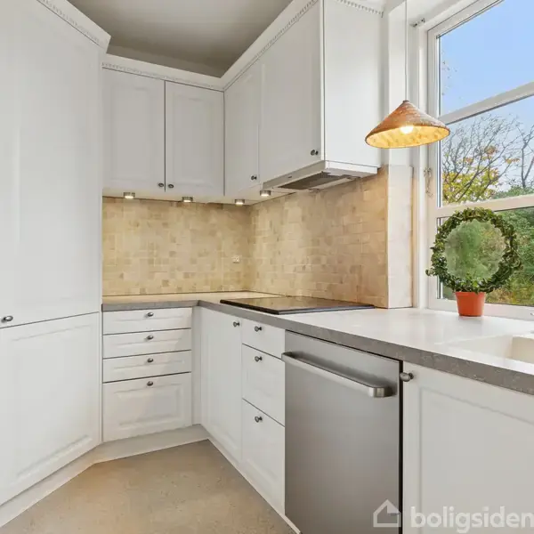 Et moderne, hvidt køkken med skabe og skuffer omkranser et hjørne. En bordplade indeholder en vask og opvaskemaskine. Store vinduer giver udsigt til træer, og to hængelamper lyser rummet op.