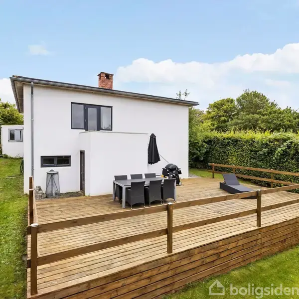 Hus med hvid facade med kvadratisk træterrasse, indeholdende havemøbler og grill, beliggende i en grøn have omkranset af buske. Klar himmel ses i baggrunden.