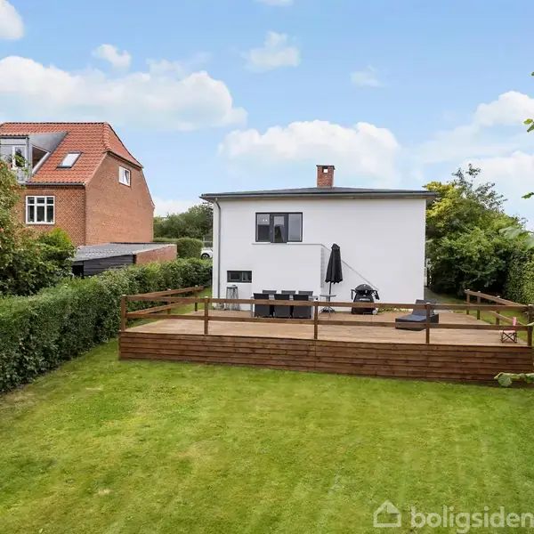 En hvid bygning står stille med en stor træterrasse foran, udstyret med udendørs møbler og en parasol. Omgivet af en grøn have med hække og et nabohus i baggrunden.