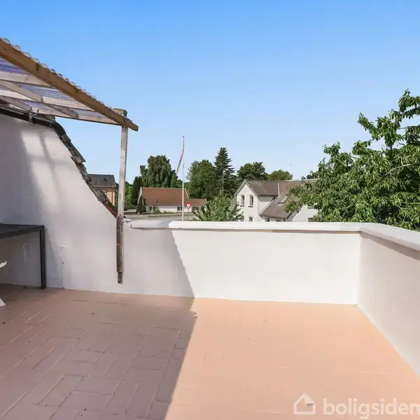 Overdækket terrasse med et lille bord og en stol, omgivet af hvidmalede vægge. Solrigt vejr med udsigt til grønne træer og nabohuse i baggrunden.