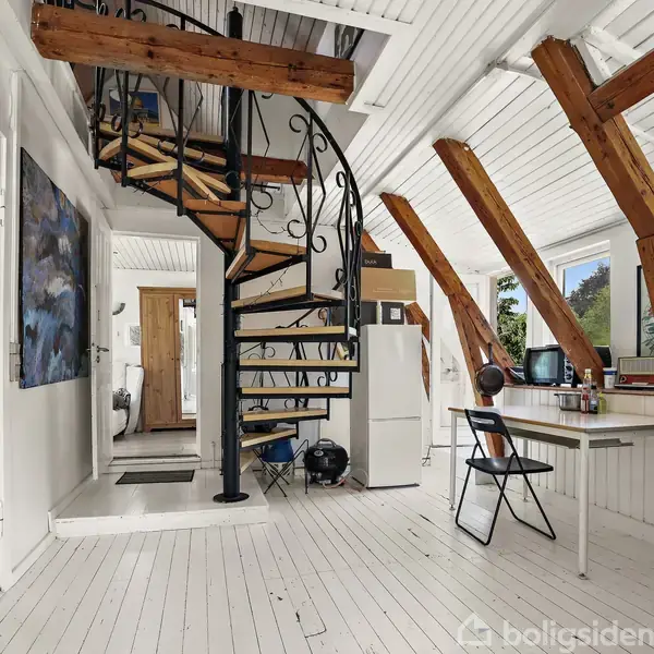 Trætrappe snor sig opad til en øvre etage i en hvidmalet stue. Store skrå vinduer slipper lys ind, mens et skrivebord og stol står ved siden af trappen.