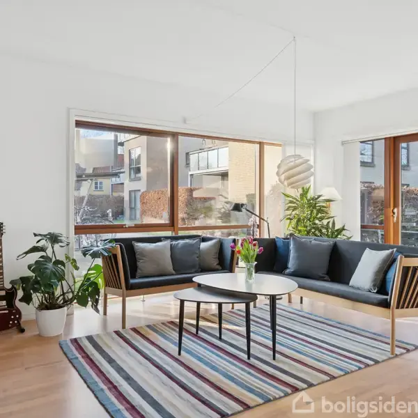 Stue med blå sofa omkring rundt bord, omgivet af indendørs planter og en stueguitar. Store vinduer med udsigt til bymiljø tillader naturligt lys, mens en stribet tæppe pynter gulvet.