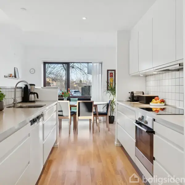 Lyst køkken med hvide skabe og bordplader i stål. Bordpladerne er dekoreret med skåle og planter. Et spisebord med stole ses ved et vindue i baggrunden, der giver naturligt lys.