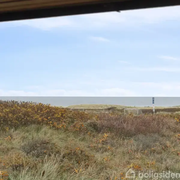 Det åbne vindue viser udsigt over en strand med høje klitter og kratbevoksning i forgrunden, rolige bølger i baggrunden og en klar blå himmel ovenover.
