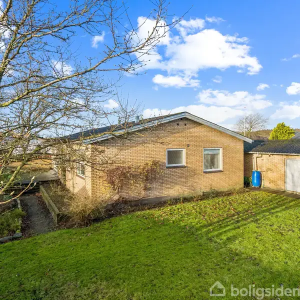 Et murstenshus står stille i en have med grøn græsplæne. Træer uden blade omgiver huset, og himlen er blå med spredte skyer.