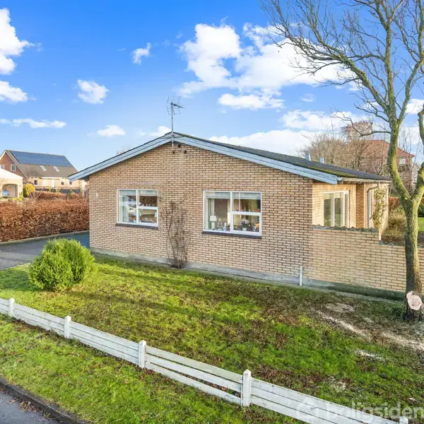 En murstensbungalow med vinduer, stående på en græsplæne, omgivet af en lav hvid træhegn. Stien ved siden fører op til huset. Baggrunden har flere huse og træer under en blå himmel.