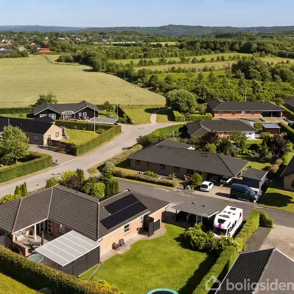 Huse med mørkt tag står stille i en beboelsesområde med grønne haver, omkranset af veje. En hvid varevogn holder parkeret ved et hus. Landskabet i baggrunden har marker og skovområder.