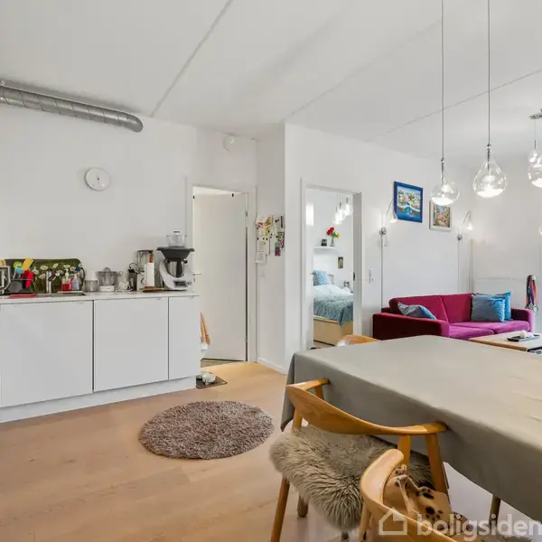 Et moderne køkkenalrum med hvide skabe, bordplade og diverse køkkenudstyr. Langbordet er dækket, omgivet af stole. I baggrunden ses en stue med sofa og store vinduer, der giver lys.