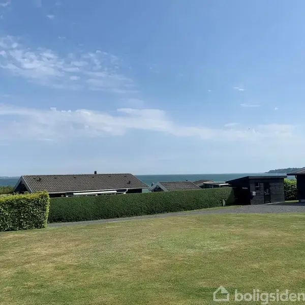 Et græsområde strækker sig foran et sort sommerhus, mens havet ses i baggrunden under en blå himmel. Adskillige småhuse ligger langs horisonten.