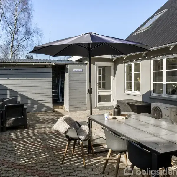 Havebord med enkelt service og stole står under en stor parasol. Solbeskinnet gårdhave med flisebelægning foran et gråt hus med vinduer og skur i baggrunden.