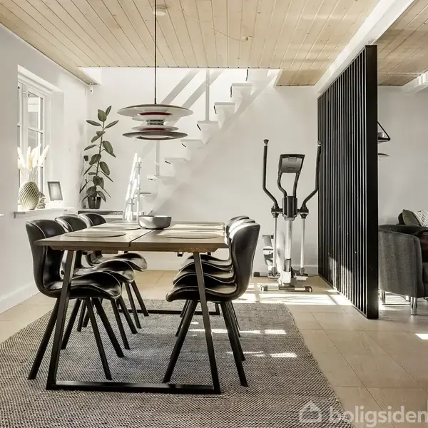 Spisebord med sorte stole står på et tæppe i en lys spisestue. En trappe fører opad, mens en motionsmaskine står til højre ved en skillevæg.