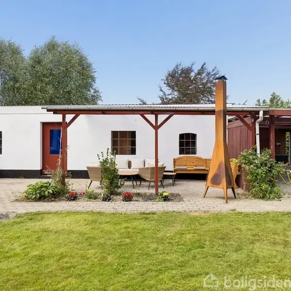 Et lille, hvidt hus med røde døre og vinduer står i en have. Foran huset er der en terrasse med havemøbler og en fritstående pejs. Grønt græs omgiver området.