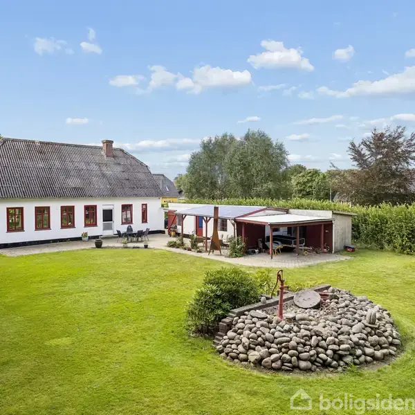 Hus med hvid facade og gråt tag står stille ved siden af carport i en velplejet have med stenbed. Baggrunden består af grønne træer og blå himmel med hvide skyer.