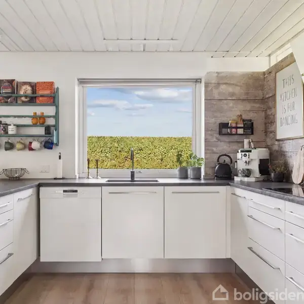 Et lyst køkken har hvide skabe og sorte bordplader, udsigt til en have gennem et stort vindue, og der er en hylde med kopper. Brættet på væggen siger: "This kitchen is for dancing."